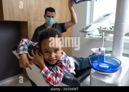 Dark-skinned child lying in a dental chair at a clinic. Child healthcare and pediatric dentistry themes with a friendly, approachable atmosphere. Indoor dental office setting suitable for healthcare communications. Stock Photo