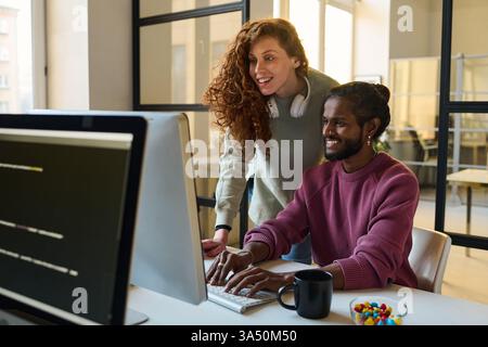 Indian male programmer sitting and working on computer near caucasian female colleague standing behind him at table in office Stock Photo