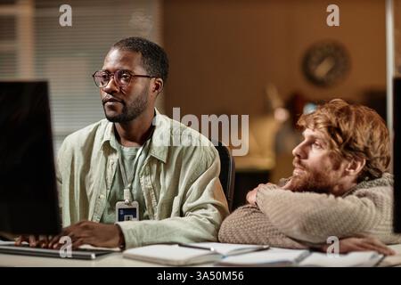Portrait of a male programmer wearing glasses working in a dimly lit office at night, with a colleague looking on. The scene emphasizes focus, collaboration and technology in a professional IT environment. Great for stories on software development, coding and cybersecurity teams. Stock Photo