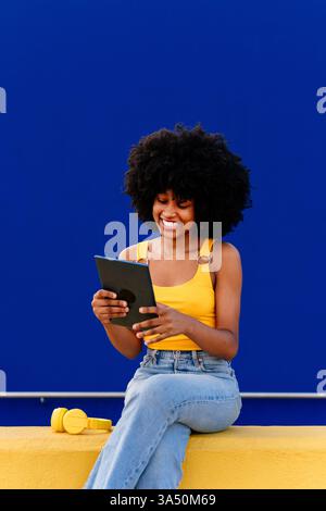 Smiling Black woman with an Afro using a tablet while sitting cross legged on a yellow fence beside a blue wall in an urban street setting. This lifestyle image reflects tech use and contemporary style. Stock Photo