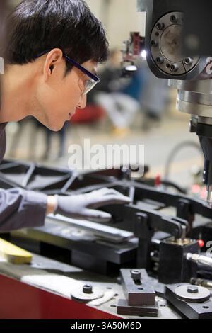 Chinese male mechanical engineer in safety glasses operates machinery in a factory setting. Ideal for engineering, manufacturing and industrial workplace visuals. Stock Photo