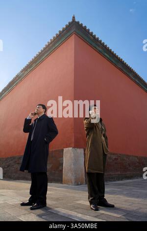 Two Chinese businesspeople stand on a sunlit street, one on a mobile phone, looking away. The backdrop blends historic architecture with modern city life in Beijing. Ideal for stories on business networking, urban commerce, and the integration of tradition with modern enterprise. Stock Photo