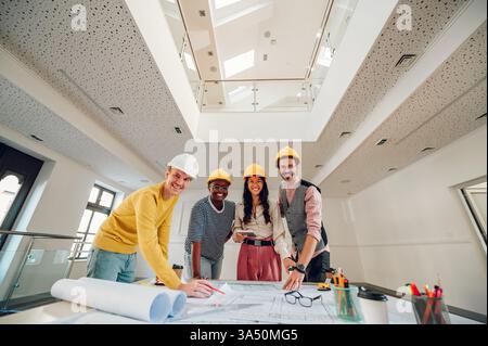 Cheerful, diverse group of architects and engineers in a modern office smile for the camera. They stand beside blueprints and a tablet, showcasing teamwork on a building project. Perfect for stories about design firms, inclusive offices, and architectural collaboration. Stock Photo