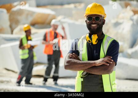 Black male engineer on a construction site wearing a safety helmet and sunglasses with arms crossed. This outdoor industrial setting highlights safety, expertise, and a professional approach to project work. Stock Photo