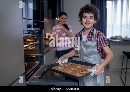 Smiling schoolboy stands beside an open oven with a cookie sheet, while a joyful woman looks on from behind. Great for home baking, family life and kitchen lifestyle themes. Conveys warmth, cheer and domestic achievement. Stock Photo