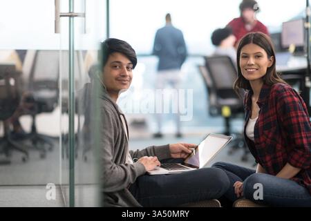 Smiling Mixed race man with earphones using laptop with caucasian female coworker sitting on floor in office Stock Photo