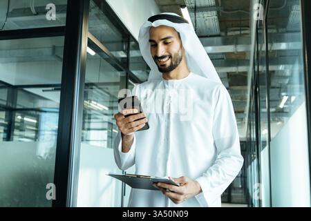 Smiling Arab man uses a smartphone while holding a folder in a corporate office hallway during daytime. This professional lifestyle image works well for business, multicultural workplaces and urban work life features. Stock Photo