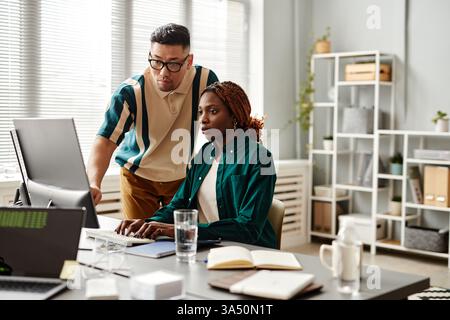 Serious Asian male programmer wearing eyeglasses standing behind Black female colleague with braids working on computer sitting at table with noteb... Stock Photo