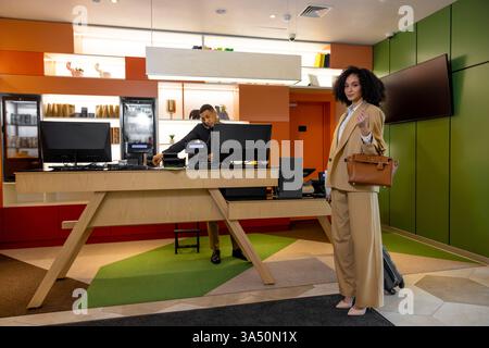 Smiling Black businesswoman with a bag and luggage at a hotel reception with a receptionist nearby. This scene is suitable for travel, business trips, hospitality services, and customer service themes. Stock Photo