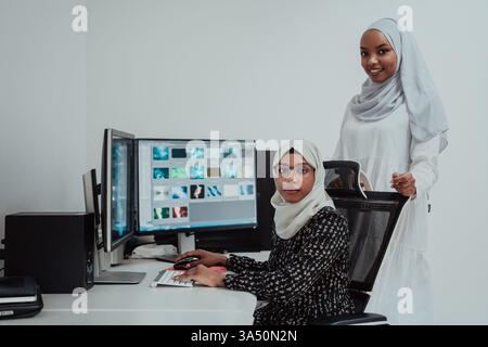 Two young Afro-American Muslim women in scarfs work together in a bright, modern office with a large screen. This scene highlights teamwork and professional collaboration in a contemporary workplace. Stock Photo