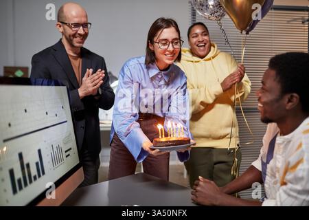 Diverse group of colleagues applaud in the office as they present a surprise birthday cake to a smiling coworker. The scene emphasizes teamwork, celebration and company culture. Stock Photo