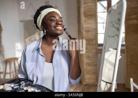African woman near a mirror applying makeup. Beauty and makeup lifestyle image suitable for cosmetics, skincare, and feminine care campaigns. Indoor, close-up vibe with a confident, polished look. Stock Photo
