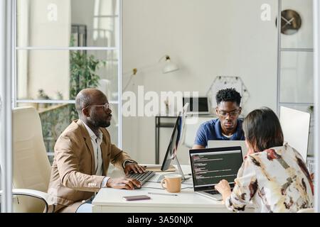 Group of diverse programmers collaborating at a table, working on new software on laptops. Perfect for technology, software development, IT, and corporate teamwork campaigns. Stock Photo