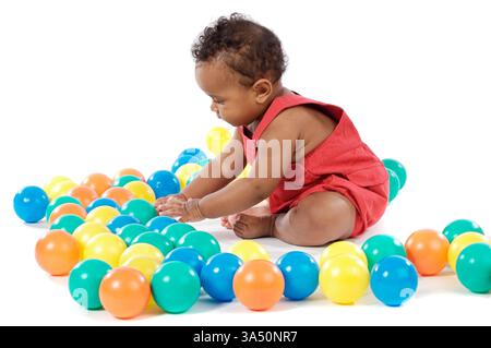 Black baby girl playing with colored plastic balls while sitting against white background Stock Photo