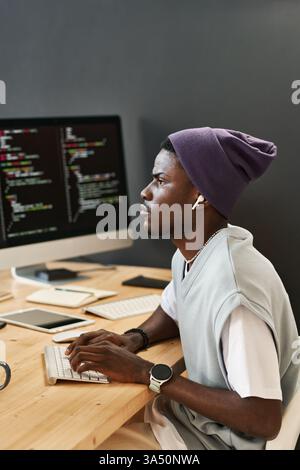 Young IT engineer at a modern workplace wears earphones and types on a keyboard while glancing at a coding screen. This image communicates focus, technology, and professional software development. Stock Photo