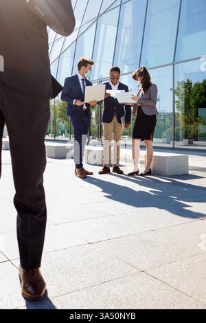 Young African American man with colleagues wearing protective masks works on a laptop in a bright office. A modern business scene emphasizing teamwork and the new normal in the workplace. Stock Photo