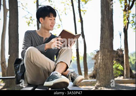 Smart and relaxed young Asian male college student reads a book while lounging in the park on the weekend. A friendly campus lifestyle image perfect for study, leisure, and outdoor living. Ideal for student life, education, and recreational outdoor content. Stock Photo