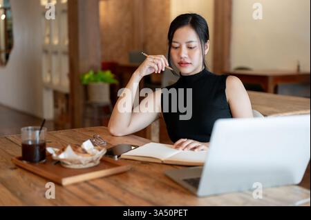 Modern millennial Asian woman reads a book while working remotely in a stylish cafe. This city lifestyle moment is ideal for features on remote work, freelance professionals, and cafe culture. Stock Photo