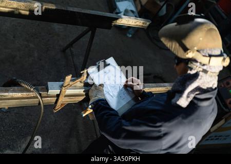 A male construction worker in a hard hat and overalls takes notes at a table in a bright garage workshop. The scene emphasizes blue collar labor, focus and industrial work. Great for topics on construction, engineering and workplace safety. Stock Photo