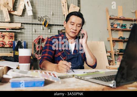 Portrait of a mature carpenter at his workshop desk, focused on a phone call with a customer while taking notes in a planner. This scene highlights skilled carpentry, woodwork and small business activity. Stock Photo