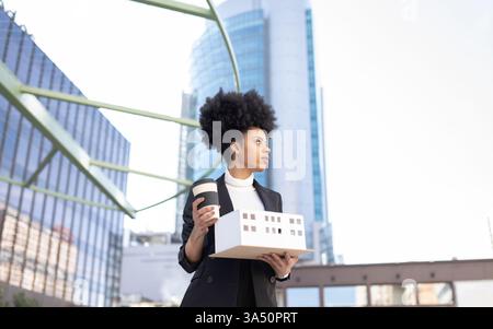 Low-angle shot of a cheerful African American female architect with a takeaway coffee beside a miniature house model. This modern, professional image suits architecture, urban design, and real estate materials. Great for project showcases and career-focused campaigns. Stock Photo