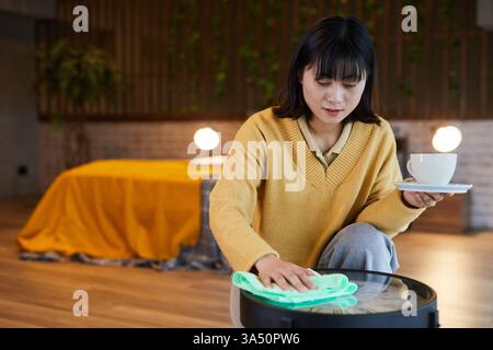 Portrait of a young Asian woman cleaning a coffee table in a bright home interior. The shot conveys domestic life with copy space for design use. Great for home lifestyle, cleaning, and family-friendly imagery. Stock Photo