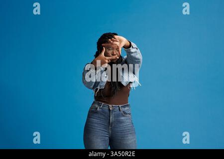 Happy Black woman with braids wearing a denim jacket smiles while framing her face with her hands against a blue studio backdrop. This contemporary portrait offers copy space for branding or advertising and works well for fashion and lifestyle themes. Stock Photo
