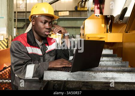 Young worker in an industrial plant stands in a workshop, using a laptop to analyze data. The focused scene works for manufacturing, engineering and technology contexts. Stock Photo