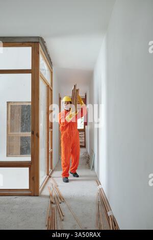 A young Vietnamese worker walks along a house corridor carrying heavy wooden building materials. The scene captures active construction and home improvement work, with emphasis on physical effort and industry. Stock Photo