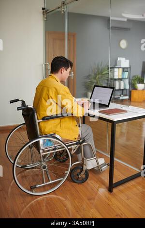 A young Vietnamese man with a disability codes on a laptop in a modern office. The inclusive workspace emphasizes accessibility and tech talent in a contemporary environment. Stock Photo