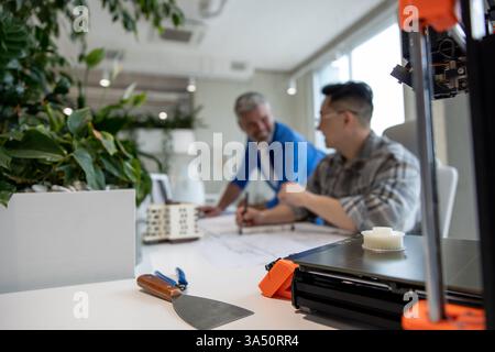 Team of two architects using a 3D printer in a bright office as a house projection and blueprints are shown. This design-focused image suits topics on architecture, 3D printing, and collaborative professional work. Stock Photo