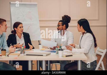 Diverse group of young colleagues discuss company projects in a boardroom during a meeting. The team collaborates around a table with a flipchart and charts visible. This scene captures teamwork, brainstorming, and a professional work environment. Stock Photo
