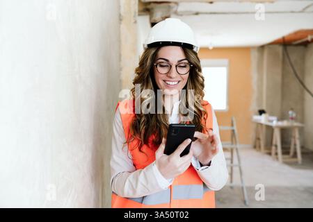 Professional architect on a construction site uses a mobile phone beside blueprints during a home renovation project. The image highlights planning, on-site coordination, and a confident forewoman wearing protective helmet and jacket. Ideal for real estate, architecture, and construction management campaigns. Stock Photo