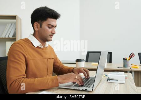 Asian man working on a laptop at a table in an office. A focused view of coding and IT work for developers and programmers. Great for technology, software development, and modern workplace visuals. Stock Photo