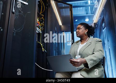 Low-angle view of a female IT engineer using a laptop in a data center, setting up a server network with copy space. This tech-forward image suits IT careers, data center marketing, and STEM promotion. Ideal for technology, networking, and enterprise infrastructure campaigns. Stock Photo