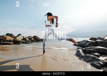 Activewear sportswoman in white runs along a sandy beach with the sea in view. This outdoors fitness shot captures energy and health in a beach setting. Perfect for fitness, wellness, or travel lifestyle campaigns. Stock Photo