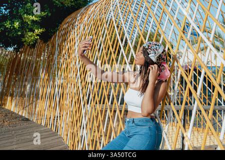 Black female in headphones taking a selfie leaning and standing against a metal arc. This candid lifestyle shot highlights music, social media and urban moments. Perfect for lifestyle, entertainment and fashion campaigns. Stock Photo