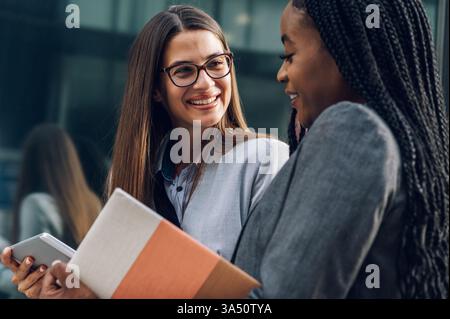 Smiling Black businesswoman with braids holding book talking to caucasian female colleague showing tablet standing outside office building during d... Stock Photo
