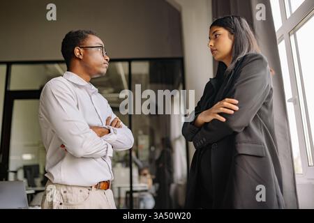Two professionals, a man and a woman, stand in a modern office with arms crossed, deep in conversation. The scene conveys tension and professionalism, suitable for business, leadership, and workplace discussions. Stock Photo