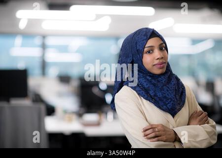 Portrait of a young Muslim African American female software developer with a blue scarf in a modern startup office. Her confident stance and contemporary workspace make this image suitable for diversity and technology themes. Great for features on leadership, coding, and inclusive workplaces. Stock Photo