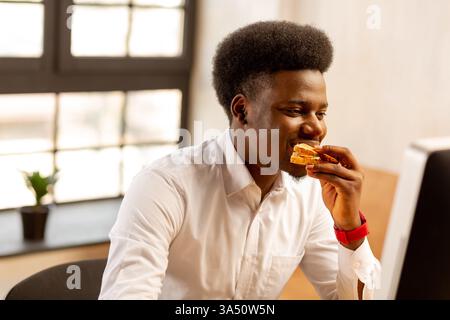Black male office worker in his 20s sits at a desk eating a sandwich while working. The modern indoor setting emphasizes focus and productivity. Great for corporate, workplace and everyday lunch break themes. Stock Photo
