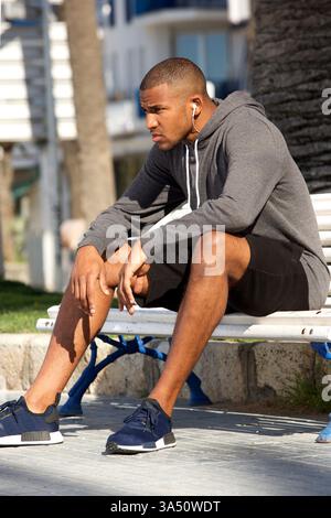 Portrait of an African American man in athletic wear resting on a bench outdoors. Suitable for urban fitness, wellbeing and athlete lifestyle visuals. Stock Photo