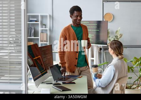 Positive colleagues share ideas during a coffee break, discussing project details in a modern office. This diverse team image works well for teamwork, collaboration, and workplace culture visuals. It conveys friendly communication and productive business environments. Stock Photo