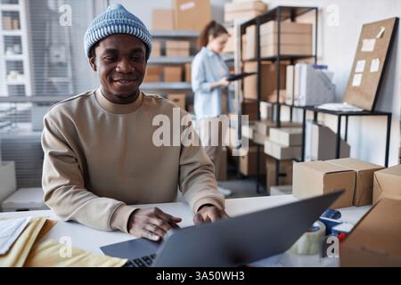 Young African American man in casual wear sits at a workstation with a laptop, taking online orders in a storage room. The scene highlights e-commerce management, digital sales, and warehouse operations. Ideal for stock imagery about online shopping, order processing, and inventory handling. Stock Photo
