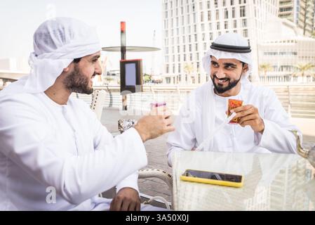 Smiling Middle Eastern men share tea and shisha at an outdoor restaurant in a Gulf city during daytime. They sit at a table, chat, and bond over a relaxed conversation as the shisha wafts in the open air. Ideal for topics on Middle East culture, friendship, and casual business networking in Dubai or the Emirates. Stock Photo