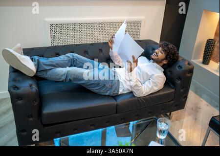 A smiling young Asian man lies on a leather sofa, reviewing a pile of documents. This lifestyle image captures a focused, casual work moment in a home or office setting. Ideal for business, productivity, or work from home campaigns. Stock Photo