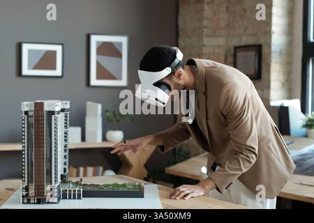 Multiracial man in the office wearing a VR headset, examining a 3D house model. A futuristic technology and design scene highlighting virtual reality in architecture and real estate. Ideal for innovation, construction tech, and 3D modelling features. Stock Photo