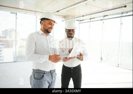 Multiracial group of male engineers wearing hard hats discuss a project on a construction site, with laptops and blueprints in view. The scene emphasizes teamwork, construction, and engineering collaboration. Ideal for corporate, industry and technology themed campaigns. Stock Photo