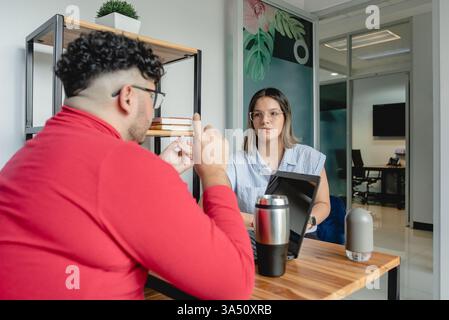Hispanic man discussing work to female colleague sitting at table with laptop in office Stock Photo