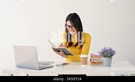 Cheerful Asian college student wearing glasses sits at her desk taking notes in a notebook. She is set against a clean white background, suitable for education and university themes. Great for student life, study, and academic materials. Stock Photo